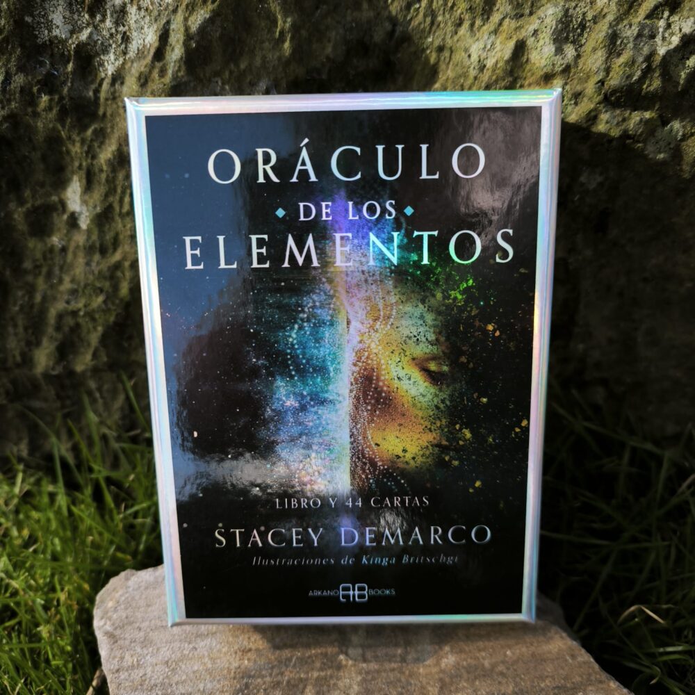 Caja del Oráculo de los Elementos con detalles metalizados e ilustración de un rostro místico en un fondo natural de bosque y piedra